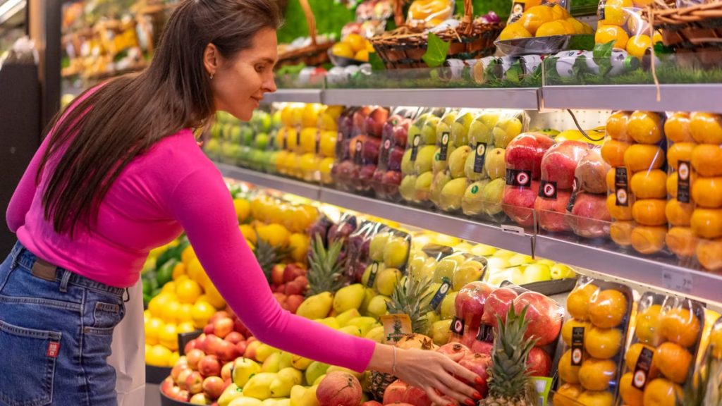 A woman in a bright pink top selecting fresh fruits in a grocery store produce aisle.