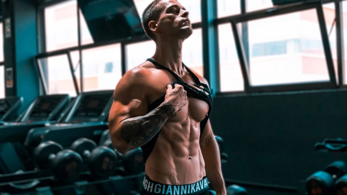 Muscular man showing abs and tattoos in a gym setting.
